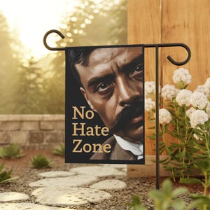 Puede incluir: Una bandera de jardín negra con las palabras "No Hate Zone" en letras doradas. La bandera presenta un retrato de un hombre con un bigote oscuro y una mirada intensa. La bandera se muestra en un soporte de metal negro en un jardín.