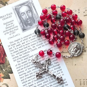 May include: A Holy Face chaplet featuring translucent red beads and matte black beads connected by silver-toned metal links. The rosary includes a silver-toned medal depicting a face and a detailed silver-toned crucifix, resting on an informational card.