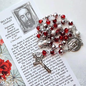 May include: A Holy Face Chaplet with red and silver beads, a crucifix, and a medal featuring a religious figure. The chaplet is accompanied by a printed explanation from the Manual of Archconfraternity of the Holy Face of 1887.