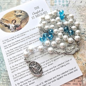 May include: A Chaplet of St. Michael prayer bead strand featuring white faux pearls and translucent blue crystal beads connected by silver-tone metal links, including a silver-tone metal medal depicting St. Michael and a prayer card.