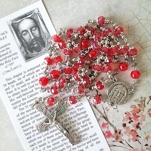 May include: Red glass beads and silver metal beads make up a rosary with a silver metal crucifix and a silver metal medallion with a depiction of Jesus' face. The text on the medallion reads "Adorina Domine Vultuor Super", which is Latin for "Worship, O Lord, Your Face Above All".