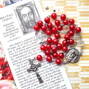 May include: A red beaded rosary with a silver crucifix and a medal featuring a religious figure. The rosary is displayed on a printed card with text about the Holy Face Chaplet.