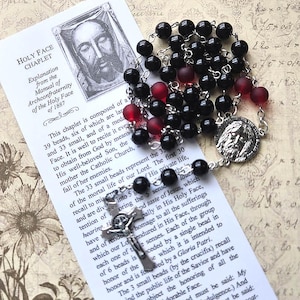 May include: A Holy Face Chaplet featuring black and matte red beads on a silver-tone metal chain. The rosary includes a circular medal depicting the face of Jesus and a silver-tone crucifix, resting on an informational card titled Holy Face Chaplet.