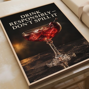 Könnte beinhalten: Ein gerahmter Wandkunstdruck mit dunklem Hintergrund und einem roten Cocktail in einem Stielglas, garniert mit einer Kirsche. Der Text DRINK RESPONSIBLY - DON'T SPILL IT ist in weißen Großbuchstaben oben auf dem rechteckigen Druck zu sehen.