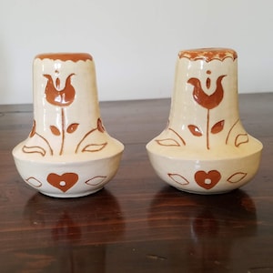 May include: A set of two ceramic salt and pepper shakers with a white glaze and brown floral designs. The shakers have a heart shape at the bottom.