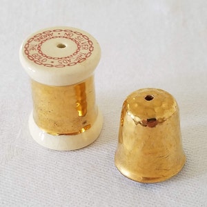 May include: A set of two gold-colored ceramic salt and pepper shakers. The salt shaker is shaped like a spool of thread and has a red and white label that reads "The Gold Ads No Silver Among The". The pepper shaker is shaped like a thimble.