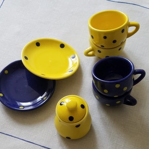 May include: A collection of ceramic tableware in yellow and navy blue, decorated with polka dots. The set includes two stacked teacups, two saucers, a small sugar bowl with a lid, and an oval serving plate. The items are arranged on a white tablecloth.