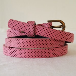 May include: A pink belt with black polka dots and a brass buckle. The belt is thin and appears to be made of a fabric material. The belt is curled up on a white surface.