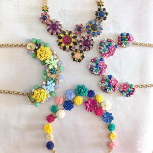 May include: A collection of colorful floral jewelry, including necklaces and earrings. The necklaces feature large, ornate flower designs with various jewel-toned accents. The earrings are round with smaller floral arrangements. Gold chains connect the pieces.