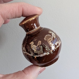 May include: A small, brown ceramic vase with a gold rim and decorative illustrations of figures playing musical instruments. The vase has a bulbous body and a narrow neck, held by a hand.