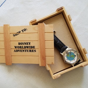 May include: A wooden crate with the text "Ship To: Disney Worldwide Adventures" on the lid. Inside the crate is a silver watch with a black leather band and a blue face with a world map design.