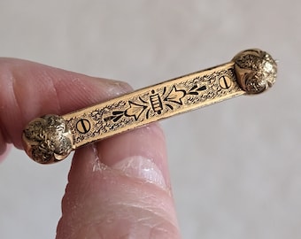 May include: A gold-toned bar brooch with an intricate black floral design. The brooch has two round, textured ends.