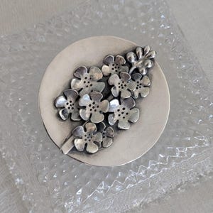 May include: A silver-toned metal brooch featuring a cluster of stylized flowers. The flowers are arranged on a circular base, with detailed petals and small, dotted centers. The brooch is likely a piece of jewelry.