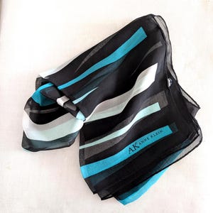 May include: Black scarf with a striped pattern in shades of blue, gray, and white. The scarf has the text "AK ANNE KLEIN" printed on it. The scarf is made of a lightweight material.