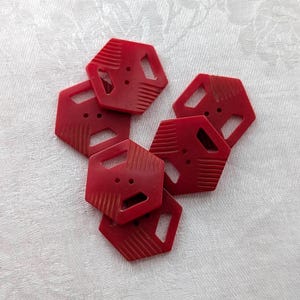 May include: A collection of six red hexagonal buttons, each featuring two central holes for attachment and decorative grooved lines along two of the outer edges. The buttons are arranged in a cluster, showcasing their uniform shape and color.
