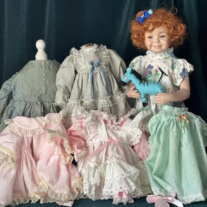 May include: A porcelain doll with red curly hair wearing a white dress with a blue and white striped dress underneath. The doll is holding a blue stuffed zebra. There are four dresses on display, two pink, one white and one green.