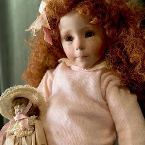 May include: A porcelain doll with red curly hair and a pink sweater holds a smaller doll. The larger doll has a delicate face with freckles and a pink bow. The smaller doll wears a dress and a bonnet. The text "DOLL KINGDOM US" is at the bottom.