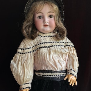 May include: A doll with brown hair and brown eyes wearing a black hat, a cream-colored blouse with black ribbon trim, and a black skirt. The doll has a pale complexion and is positioned against a black background with the text DOLL KINGDOM US.