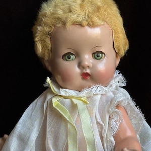 May include: Vintage doll with blonde hair and green eyes, wearing a white dress with lace trim and a yellow ribbon. The doll has rosy cheeks and red lips. The image is a close-up shot of the doll's face and upper body.