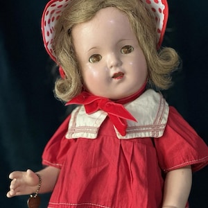 May include: A vintage doll wearing a red dress with white stitching and a white collar. The doll has blonde hair, brown eyes, and a red and white polka dot bonnet. A heart-shaped charm hangs from the doll's wrist.