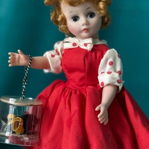 May include: A vintage doll with auburn hair, wearing a red dress with white polka dot sleeves and collar. The doll holds a clear, cylindrical container with a black and gold object inside. The doll has a pink bow in her hair.