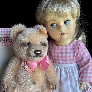 May include: A vintage teddy bear with a pink bow and a doll with blonde hair and blue eyes. The doll wears a pink and white checkered dress and a white apron. The teddy bear has a tag that says "Original Marke".