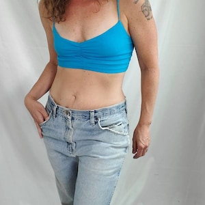 K&ouml;nnte beinhalten: Eine Frau tr&auml;gt ein hellblaues Crop-Top und eine hellblaue Jeans mit einem ausgefransten Knie. Sie hat lockiges braunes Haar und ein Tattoo auf der rechten Schulter.