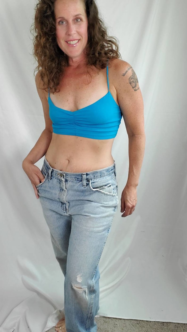 K&ouml;nnte beinhalten: Eine Frau tr&auml;gt ein hellblaues Crop-Top und eine hellblaue Jeans mit einem ausgefransten Knie. Sie hat lockiges braunes Haar und ein Tattoo auf der rechten Schulter.