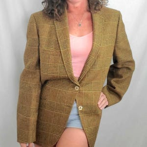 May include: A brown plaid blazer with a single gold button closure. The blazer has a classic design with a notched lapel and long sleeves. The plaid pattern features black and yellow lines. The blazer is worn over a pink top.