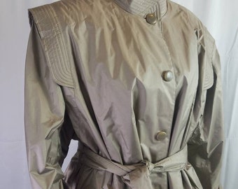 Op de afbeelding: Een beige trenchcoat met een hoge kraag en knoopsluiting. De jas heeft schoudervullingen, een ceintuur in de taille en zakken. De stof heeft een lichte glans en de jas wordt op een mannequin gepresenteerd.