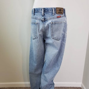 K&ouml;nnte beinhalten: Hellblaue Jeans aus Denim mit einem braunen Leder-Patch, auf dem hinten "Wrangler" steht. Die Jeans haben eine lockere Passform und ein gerades Bein.