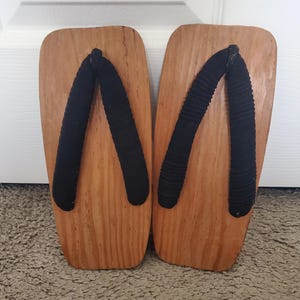 May include: A pair of geta sandals featuring rectangular wooden bases with a natural light brown wood grain finish and thick black fabric thong straps. The sandals have a flat, solid construction with rounded corners at the top and bottom edges.