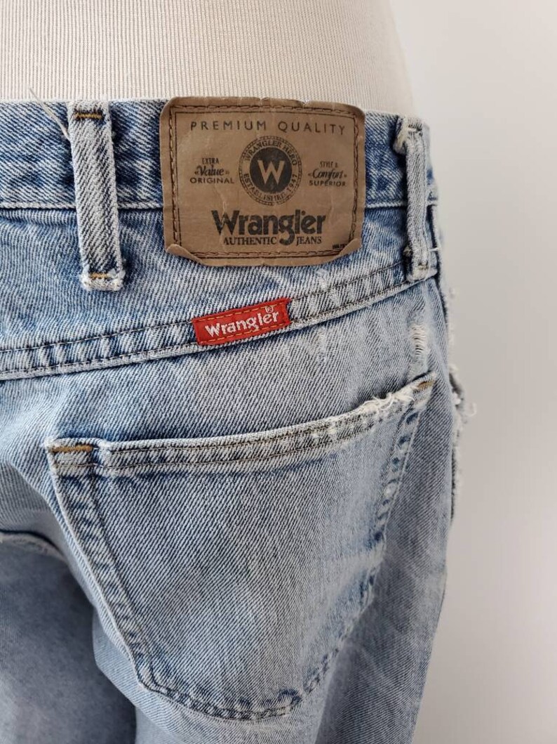 K&ouml;nnte beinhalten: Hellblaue Jeans aus Denim mit einem braunen Leder-Patch, auf dem "Wrangler Authentic Jeans" steht, und einem roten Patch mit der Aufschrift "Wrangler".