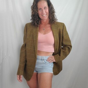 May include: A woman wearing a brown plaid blazer over a pink top and light blue denim shorts. The blazer has a classic plaid pattern in shades of brown and yellow. The outfit is casual and stylish.