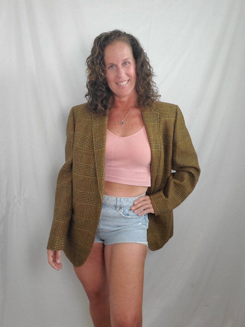 May include: A woman wearing a brown plaid blazer over a pink top and light blue denim shorts. The blazer has a classic plaid pattern in shades of brown and yellow. The outfit is casual and stylish.