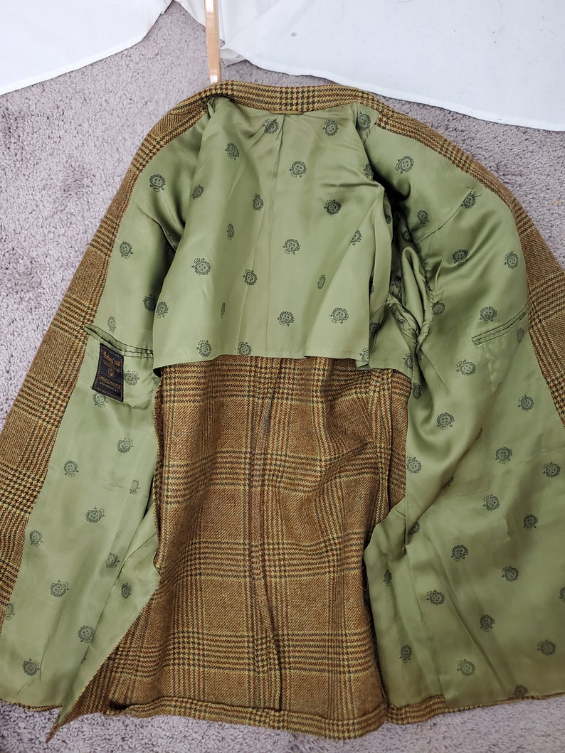 May include: A brown and tan plaid blazer with a green interior lining. The lining features a repeating pattern of small, dark green circular designs. The blazer is open, revealing the interior construction and details.