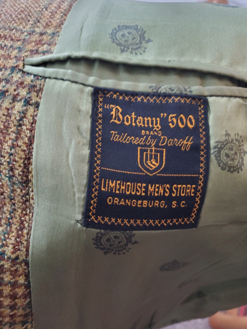 May include: Close-up of the interior of a tweed jacket. The lining is olive green with a repeating pattern. A navy blue label reads "Botany" 500 BRAND Tailored by Daroff, and "LIMEHOUSE MEN'S STORE ORANGEBURG, S.C."