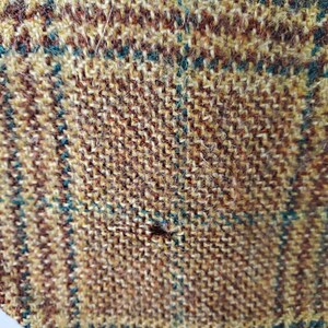 May include: Close-up of a textured fabric with a plaid pattern. The fabric features a mix of brown, tan, and teal threads woven together. The weave creates a detailed, slightly rough surface.