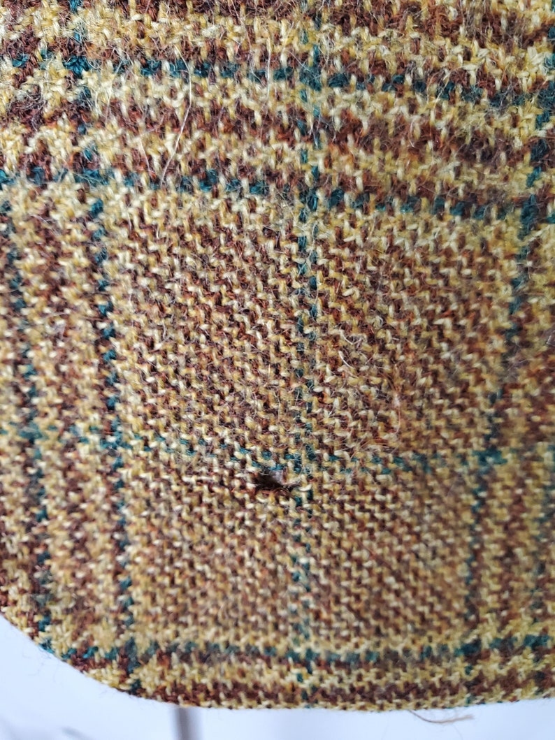 May include: Close-up of a textured fabric with a plaid pattern. The fabric features a mix of brown, tan, and teal threads woven together. The weave creates a detailed, slightly rough surface.