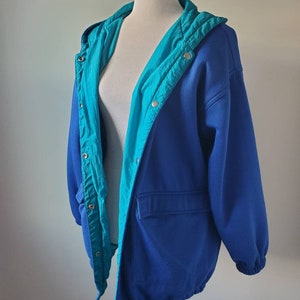 Puede incluir: Una chaqueta reversible azul y turquesa con capucha y cordón en la parte inferior. La chaqueta tiene cierre de cremallera y varios bolsillos.