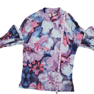 Puede incluir: Una blusa de manga larga con un estampado floral en tonos rosa, morado, azul y blanco. La blusa tiene un cuello alto y mangas acampanadas. Una cinta m&eacute;trica est&aacute; colocada sobre la blusa.
