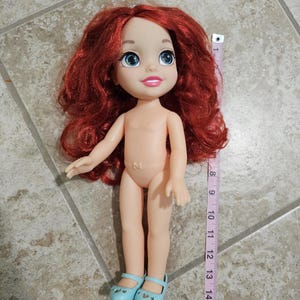 May include: A doll with red hair, blue eyes, and light blue shoes. The doll is approximately 15 inches tall. A measuring tape is next to the doll.
