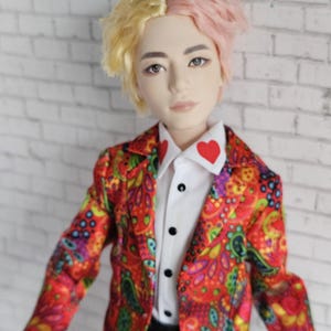 Può includere: Bambola con capelli bicolore, metà gialli e metà rosa, che indossa un blazer rosso con stampa paisley su una camicia bianca con un dettaglio a cuore rosso. La bambola ha un'espressione neutra e pantaloni neri.