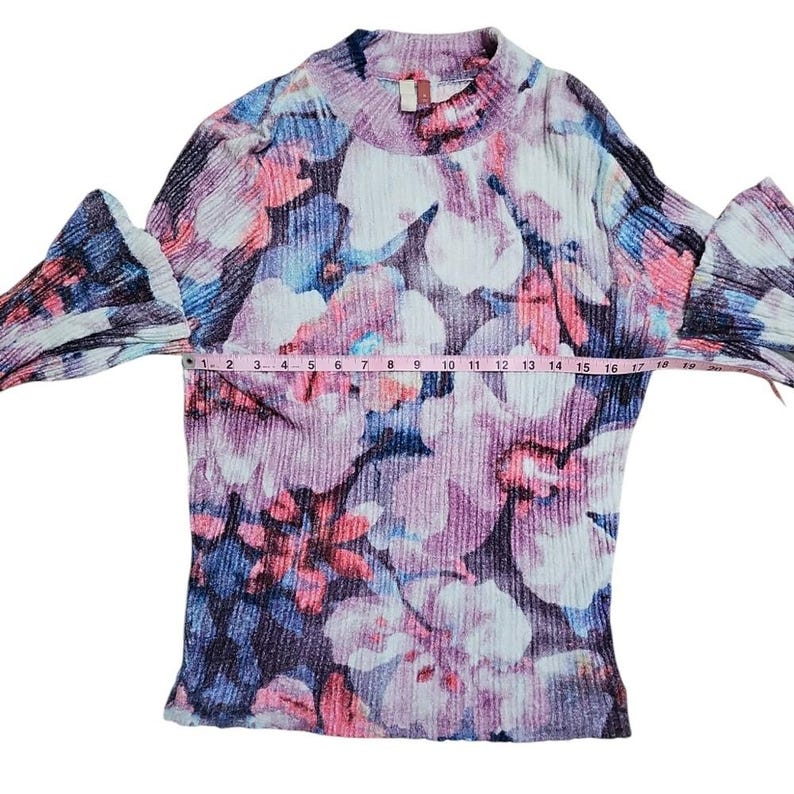 Puede incluir: Una blusa con estampado floral con cuello alto y mangas largas acampanadas. La blusa presenta un patr&oacute;n de flores rosas, moradas, azules y blancas. La manga mide aproximadamente 43 cm.