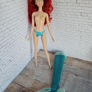 May include: A doll with red hair, blue eyes, and a turquoise bikini stands next to a detached mermaid tail. The tail is teal with a swirling pattern and a sheer, light blue fabric at the end. The doll is on a wooden surface.