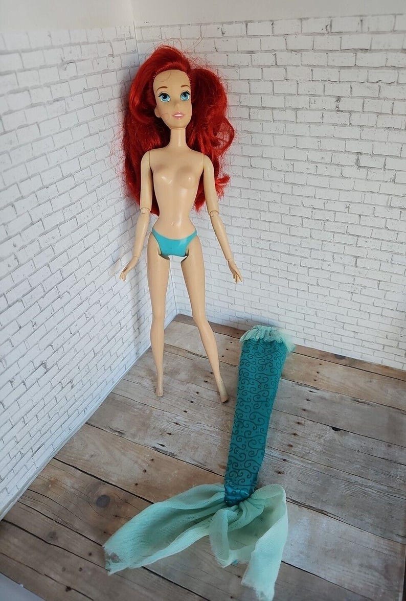May include: A doll with red hair, blue eyes, and a turquoise bikini stands next to a detached mermaid tail. The tail is teal with a swirling pattern and a sheer, light blue fabric at the end. The doll is on a wooden surface.