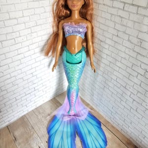 May include: A mermaid doll with long auburn hair and a fair complexion. The doll wears a purple and blue top and a teal, blue, and pink mermaid tail. The tail has large, fan-shaped fins in shades of blue and purple.