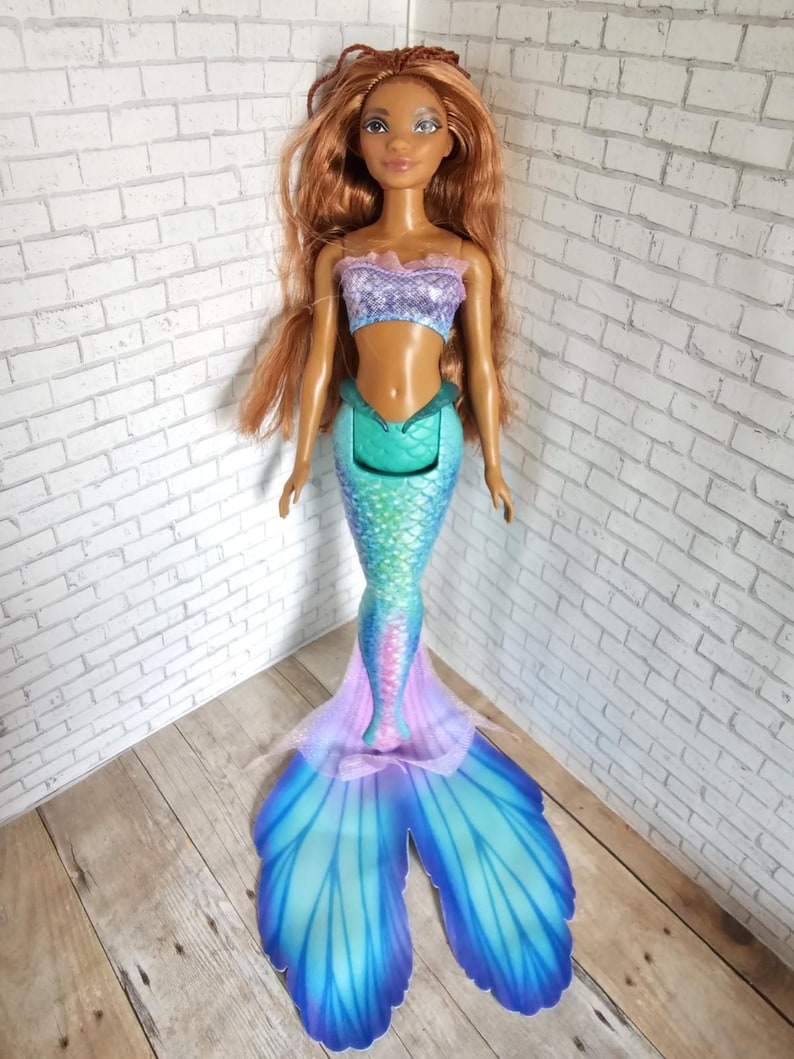 May include: A mermaid doll with long auburn hair and a fair complexion. The doll wears a purple and blue top and a teal, blue, and pink mermaid tail. The tail has large, fan-shaped fins in shades of blue and purple.