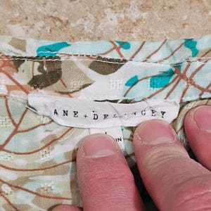 May include: Close-up of a floral patterned fabric with a tag that reads "ANE + DELANCEY". The fabric has a light blue background with brown and teal floral designs. The tag is white with black lettering.