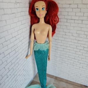 May include: A doll of a mermaid with red hair and blue eyes. The doll has a teal tail with a swirling pattern and a matching teal top. The doll is standing in front of a white brick wall.